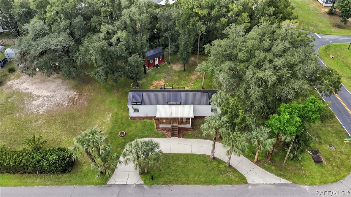 Aerial Property View  A beautiful overhead perspective of the .29-ACRE CORNER HOMESITE, showcasing the METAL ROOF, COVERED FRONT PORCH, circular concrete driveway, mature trees & detached storage building.