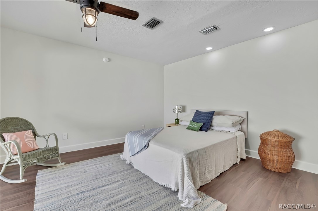 Same bedroom, other angle: bed with white bedding and a striped throw, a navy pillow, white pillows and a green "happy place" pillow, a small nightstand with a green lamp, a wicker rocking chair at the left with a pink pillow, a grey rug, dark wood-look floor, a ceiling fan with a lantern light, recessed lights, and a smoke alarm on the wall.