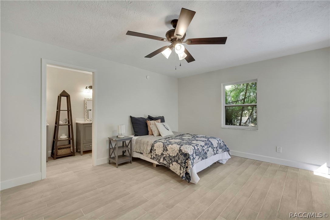Primary bedroom: bed with navy-and-white floral bedding, dark navy pillows, a patterned pillow and a white bird-print pillow, a grey nightstand with a lamp, a doorway at the left opening to a bathroom vanity area with a dark ladder shelf, a grey vanity and an ornate mirror, a window with palms at the right, light plank tile floor, and a dark ceiling fan with lights.