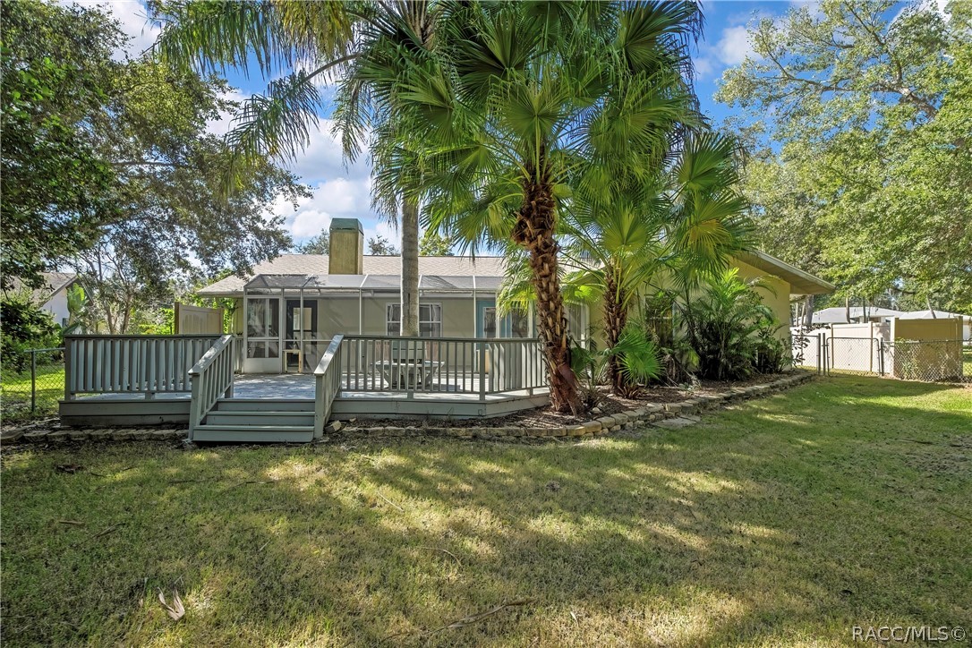 Rear of the house from the lawn: light stucco walls, a shingle roof, a stucco chimney with a metal cap, a screen enclosure with blue doors, a grey wood deck with steps and railing, palm trees, a fenced lawn in the foreground, and neighbouring houses and chain-link and vinyl fences at the sides.