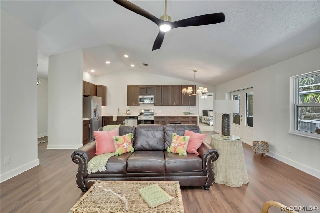 Open living room and kitchen: vaulted ceiling, black ceiling fan with light, white stone fireplace at the right with a wood mantel, mesh screen, black tile hearth and black fireplace tools, brown leather sofa with pink and patterned pillows and a green throw, a wicker trunk coffee table, a beige armchair, dark kitchen cabinets with stainless range, microwave and dishwasher, a sink with a gooseneck faucet, a dining chandelier, French doors to the porch, windows, and wood-look plank floor.
