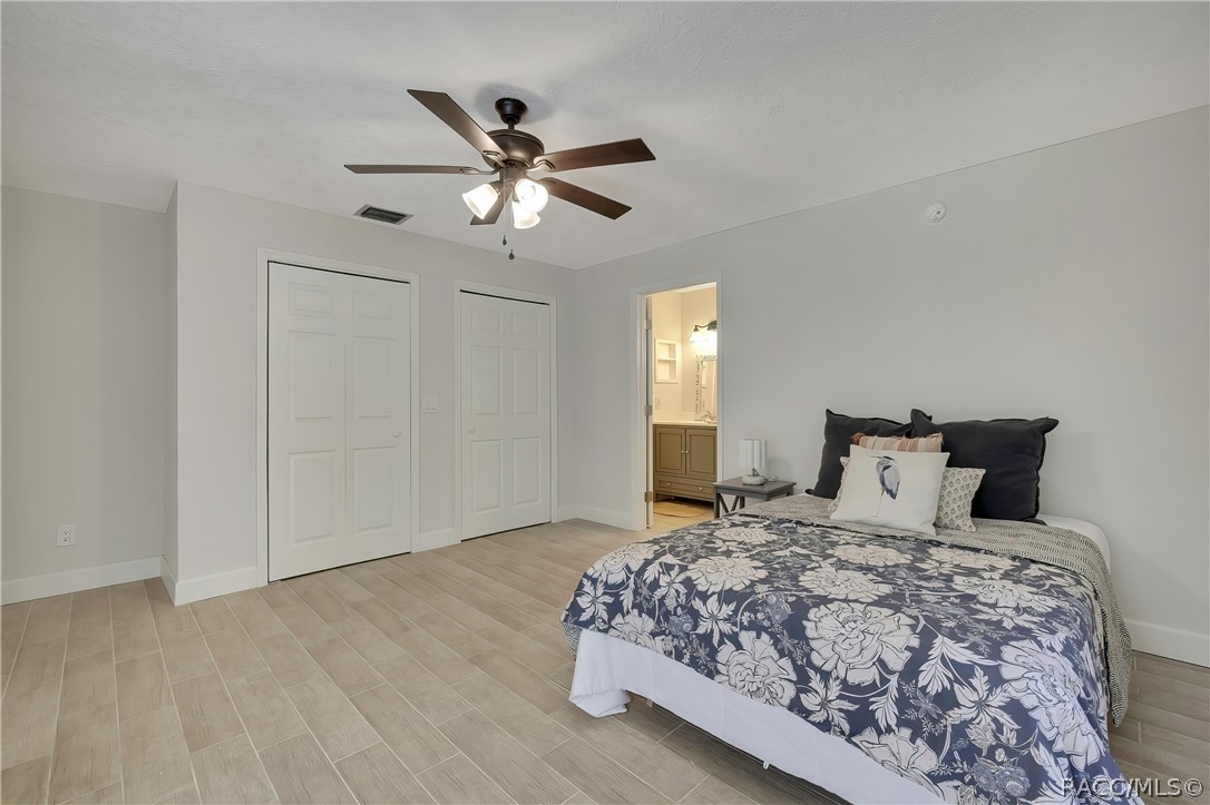 Primary bedroom, third angle: bed with navy-and-white floral bedding and a white bird-print pillow, two white closet doors at the left, a doorway in the centre opening to the bathroom vanity area, a grey nightstand with a lamp, light plank tile floor, white walls, and a dark ceiling fan with lights.