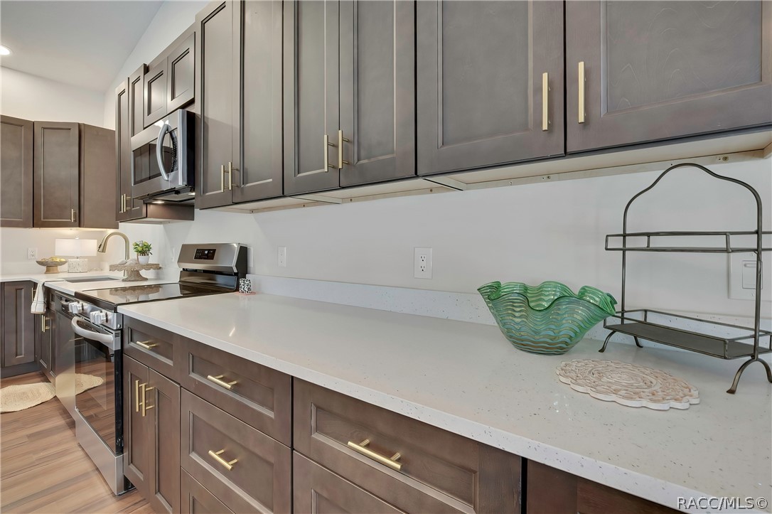 Kitchen counter run: dark cabinets with gold bar pulls, white speckled counters, an electric glass-top range and a microwave above it, a sink with a gooseneck faucet at the left, a green glass bowl, a black wire rack, a white doily placemat and a small patterned container on the counter, and wood-look plank floor.