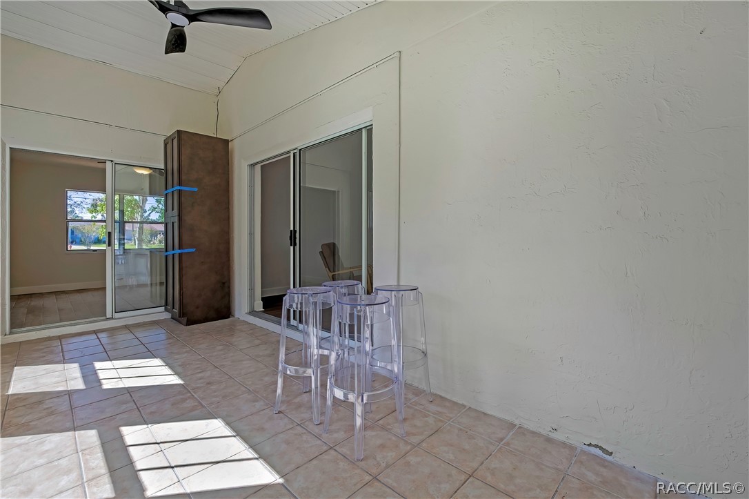 Screened porch, side angle: tiled floor, clear acrylic stools in the centre, sliding glass doors to the bedrooms, a tall dark cabinet with blue painter's tape at the left, a doorway at the left to an empty bedroom, a black ceiling fan at the top of the frame, and a white beadboard ceiling.