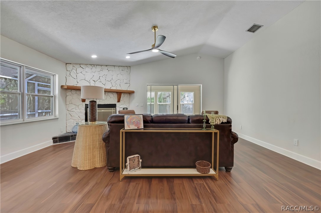 Living room seen from behind the sofa, facing the fireplace: white stone fireplace with a wood mantel, a wicker side table with a tall dark lamp, a gold console table behind the brown leather sofa with a framed picture, a small clock, a basket and green candlesticks, a green throw over the sofa, windows at the left looking onto the screened porch, sliding glass doors in the centre to the Florida room, vaulted ceiling, black ceiling fan with light, recessed lights, and wood-look plank floor.
