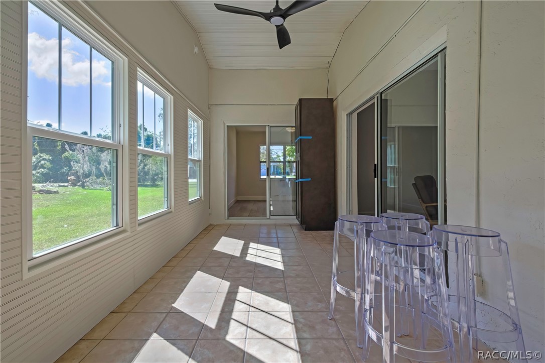 Screened porch, opposite end: tiled floor, windows at the left looking onto the lawn and trees, clear acrylic stools in the foreground at the right, sliding glass doors at the right to the bedrooms, a tall dark cabinet with blue painter's tape in the centre, a doorway at the far end to an empty bedroom, a black ceiling fan at the top of the frame, and a white beadboard ceiling.
