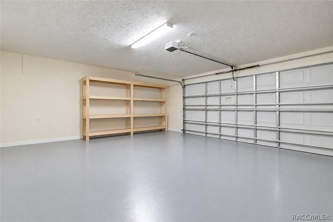 Clean, empty garage interior: smooth grey painted concrete floor, a white sectional garage door with metal rails and a garage door opener, a fluorescent tube light, a textured ceiling, beige walls, and a light wood empty shelving unit with two bays and four shelves against the back wall. No mower, boxes or clutter in this shot  this is a different, newer garage photo than the two cluttered garage shots in the old set.