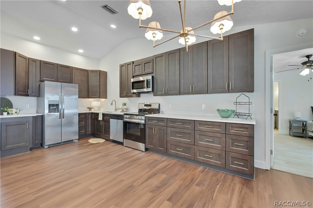 Wide kitchen view: dark grey-brown cabinets with gold bar pulls, white speckled counters, a stainless side-by-side refrigerator with a water/ice dispenser, an electric glass-top range and oven, a microwave above the range, a dishwasher, a sink with a gooseneck faucet and a towel, a small plant, a green glass bowl and a black wire rack on the counter, a chandelier at the top of the frame, recessed lights, wood-look plank floor, and a doorway at the right opening to the primary bedroom