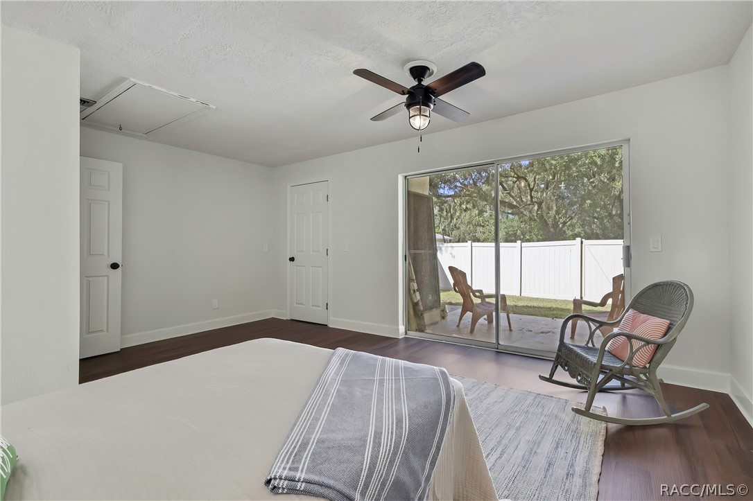 Same bedroom, third angle: bed in the foreground with white bedding, a striped throw and a green pillow at the edge, a wicker rocking chair at the right with a pink pillow, a grey rug, dark wood-look floor, sliding glass doors in the centre to a fenced patio with a white vinyl fence and lawn chairs outside, white doors at the left, an attic hatch in the ceiling, and a ceiling fan with a lantern light.