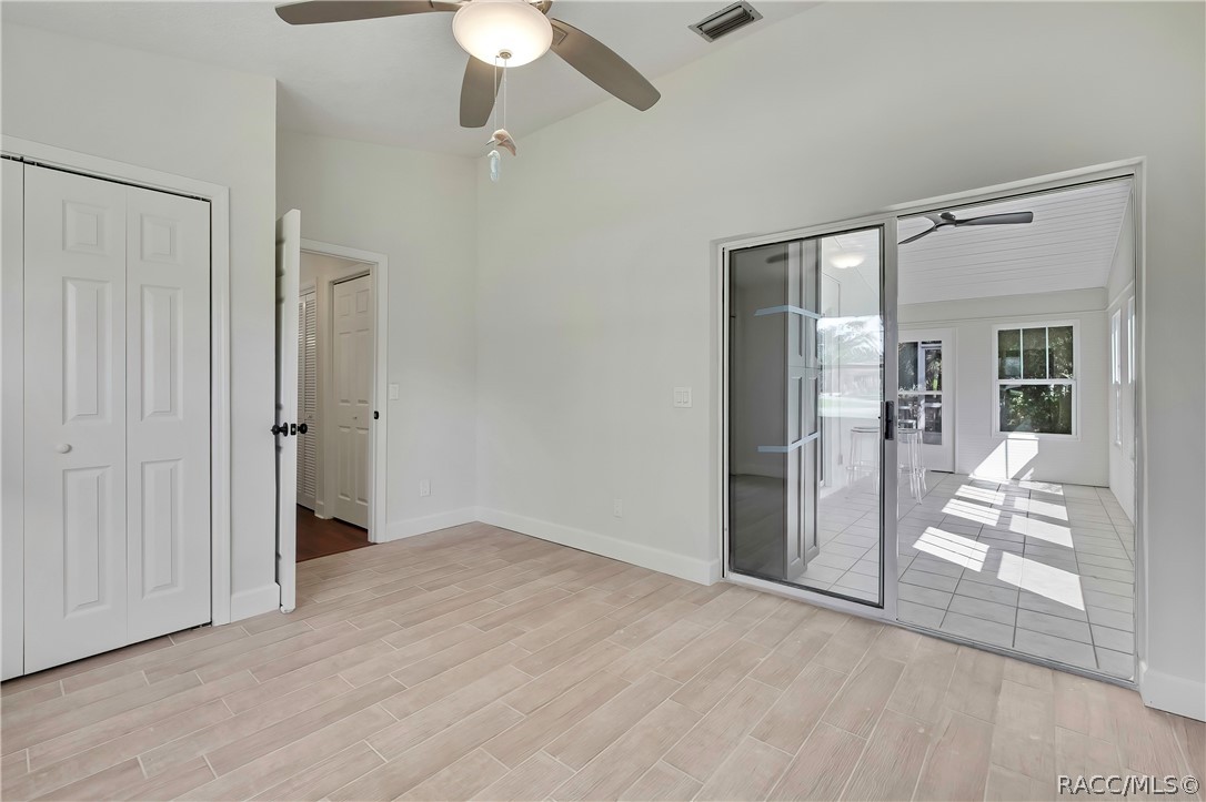 Empty bedroom: light plank tile floor, white double closet doors at the left, an open doorway to a hallway with a dark wood floor, a sliding glass door at the right to the screened porch with a tiled porch floor and chairs visible through it, a ceiling fan with a dome light, and white walls. No furniture.