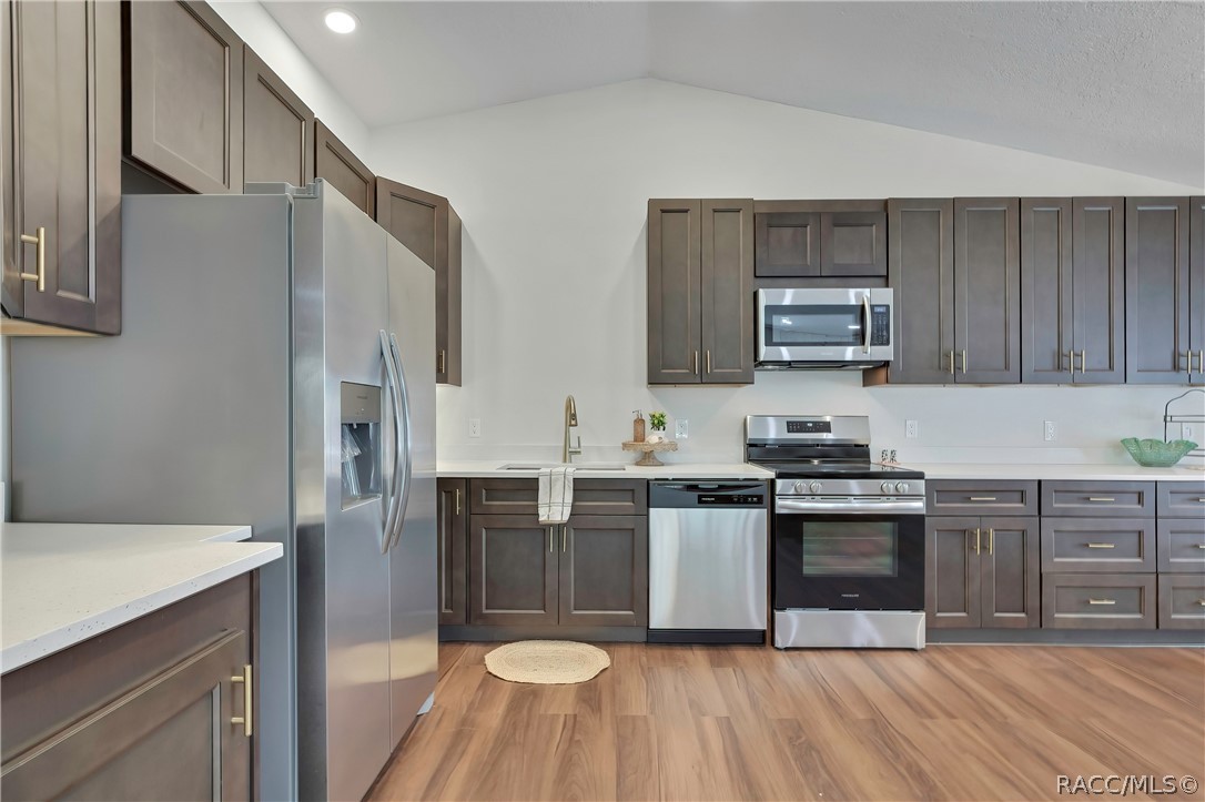Wide kitchen view: dark grey-brown cabinets with gold bar pulls, white speckled counters, a stainless side-by-side refrigerator with a water/ice dispenser, an electric glass-top range and oven, a microwave above the range, a dishwasher, a sink with a gooseneck faucet and a towel, a small plant, a green glass bowl and a black wire rack on the counter, a chandelier at the top of the frame, recessed lights, wood-look plank floor, and a doorway at the right opening to the primary bedroom.