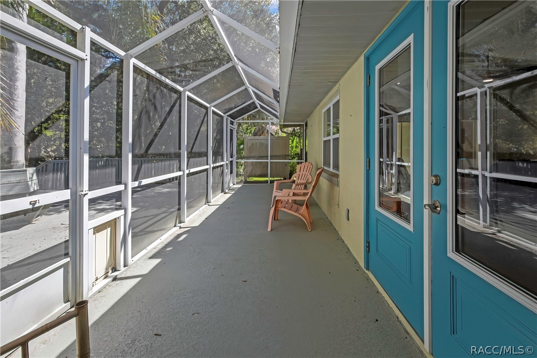 Inside the screen enclosure, along the house wall: concrete floor, white aluminium frame and screen roof, blue entry doors with glass at the right, a window, beige plastic chairs in the centre, a screen door with a pet door at the left, and a grey wood deck beyond.