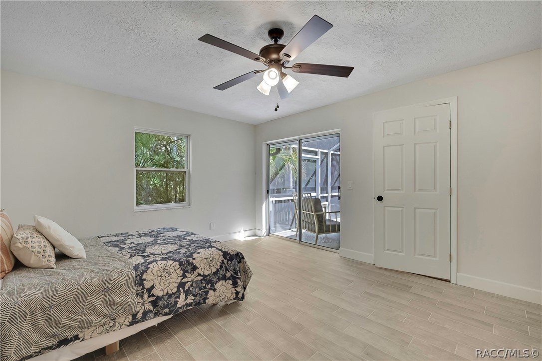 Primary bedroom from the porch side: bed with navy-and-white floral bedding, white and patterned pillows, light plank tile floor, a dark 5-blade ceiling fan with lights, a window with palms outside, a sliding glass door to the screened porch with a chair outside, a white closet door at the right, white walls and a textured ceiling.