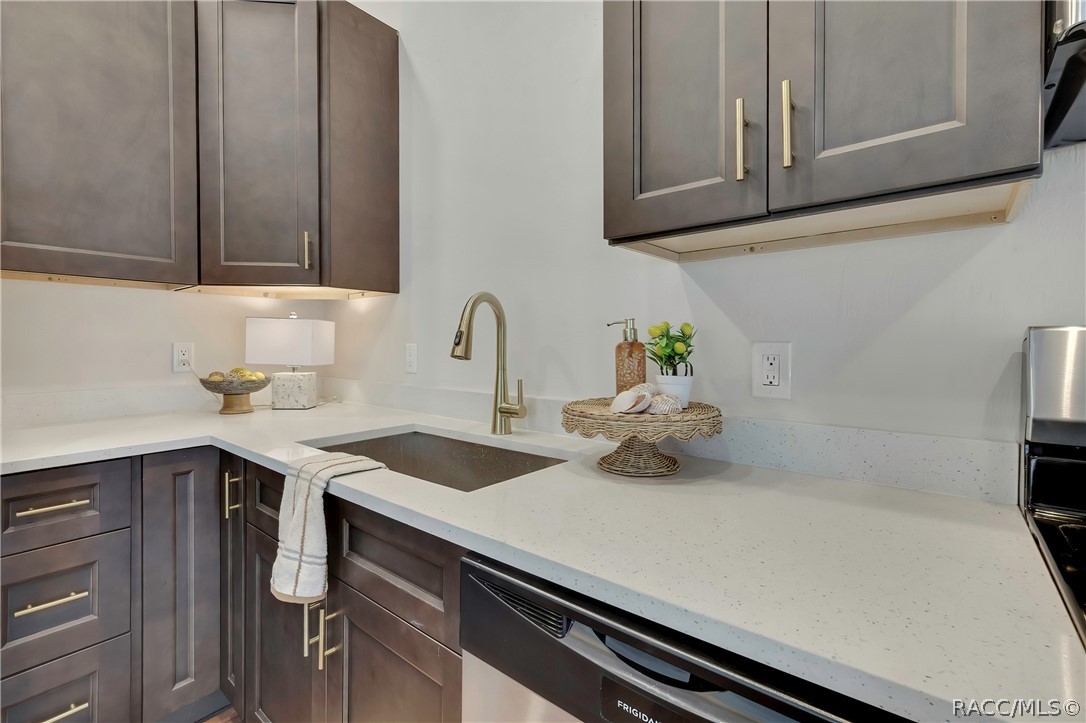 Kitchen sink close-up: brushed-gold gooseneck faucet, dark undermount sink, white speckled counters, dark cabinets with gold pulls, a striped towel over the cabinet door, a Frigidaire dishwasher at the right, the edge of the range at the far right, a wicker cake stand with shells, a soap dispenser and a small potted plant on the counter, under-cabinet lighting, and a small lamp and bowl in the corner at the left.