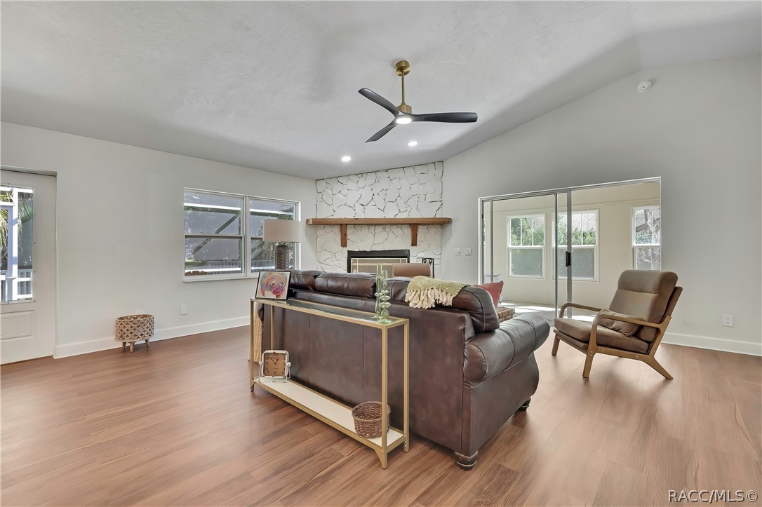Living room seen from behind the brown leather sofa: vaulted white ceiling, black 3-blade ceiling fan with brass mount and light, recessed lights, white stone fireplace with a wood mantel and black tile hearth, sliding glass doors to the screened porch / Florida room at the right, windows looking onto the porch, a gold console table behind the sofa with a framed picture, glass candlesticks, a small clock and a basket below it, a beige armchair, a green throw over the sofa, wood-look plank floor