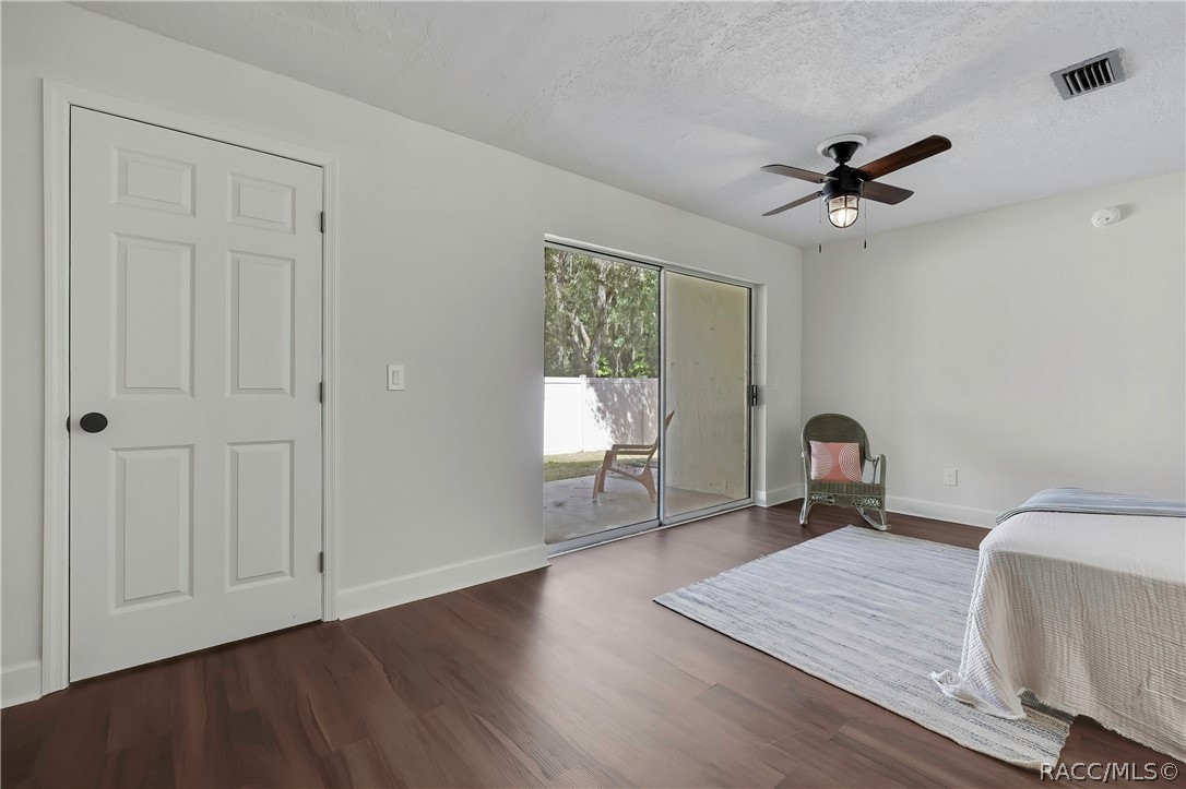 Bedroom: bed at the right with white bedding and a striped throw, a wicker rocking chair with a pink pillow, a grey rug, dark wood-look floor, a ceiling fan with a lantern light, a white door at the left, and a sliding glass door in the centre to a fenced patio with lawn chairs outside and a white vinyl fence, with trees beyond.