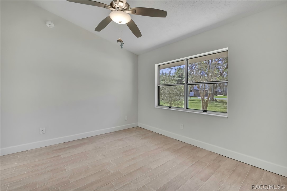 Empty bedroom: light plank tile floor, a large double window looking onto trees, a neighbouring house and lawn, a ceiling fan with a dome light, white walls, wall outlets and a smoke alarm. No furniture.