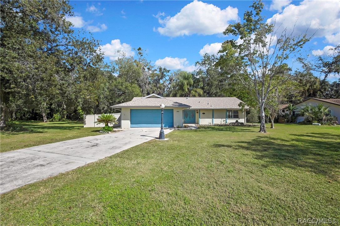 Front of the house seen from the end of the long concrete driveway: blue garage door and blue entry doors, white vinyl fence and a palm plant at the left, a black lamp post with a white globe in the centre of the lawn, green lawn, a neighbouring house at the right, trees and palms behind the house, blue sky with white clouds.