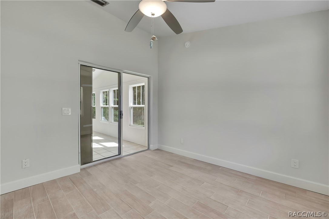 Empty bedroom: light plank tile floor, a sliding glass door at the left to the screened porch with a tiled porch floor and windows visible through it, a ceiling fan with a dome light at the top of the frame, white walls and wall outlets. No furniture.