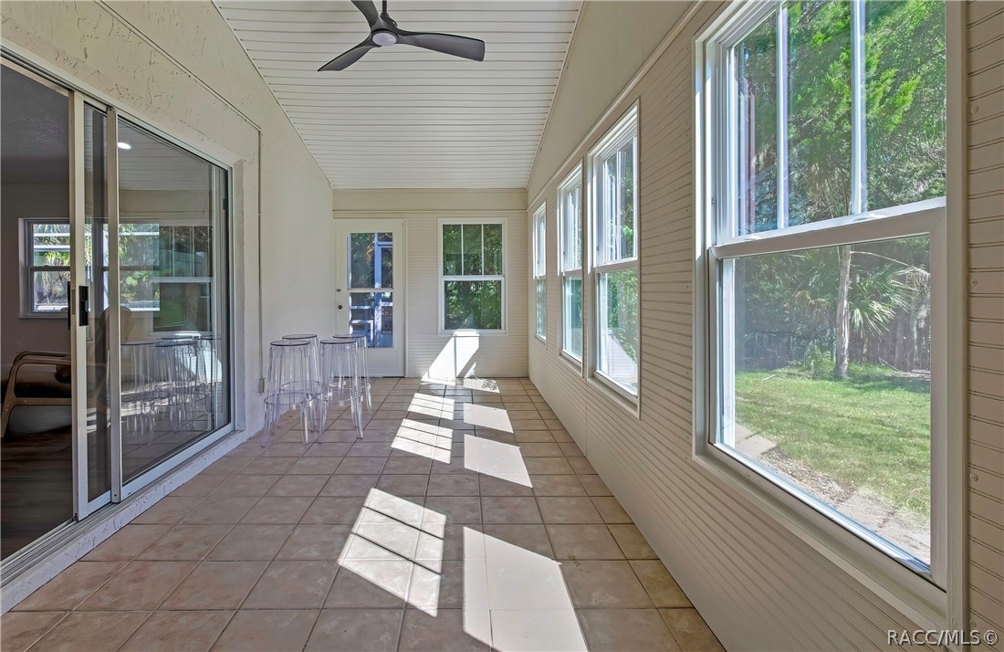 Screened porch, long view: tiled floor, white beadboard ceiling with a black ceiling fan, windows at the right looking onto the lawn and trees, sliding glass doors at the left to the living room, clear acrylic stools, and a white door and a window at the far end, with sunlight patches across the floor.