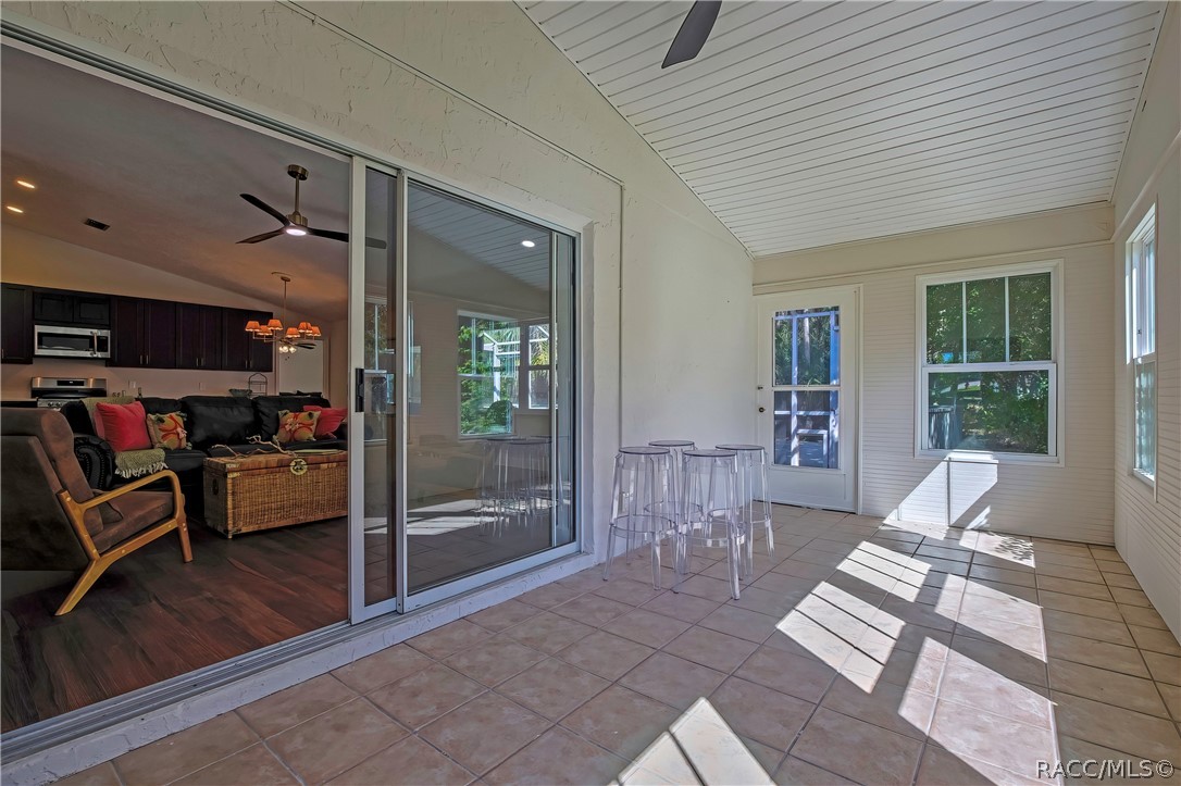 Screened porch from the other end: tiled floor, white beadboard ceiling, sliding glass doors at the left to the living room and kitchen, with the sofa, wicker trunk and armchair visible through them, clear acrylic stools in the centre, and a white door and a window at the far end, with sunlight patches across the floor.