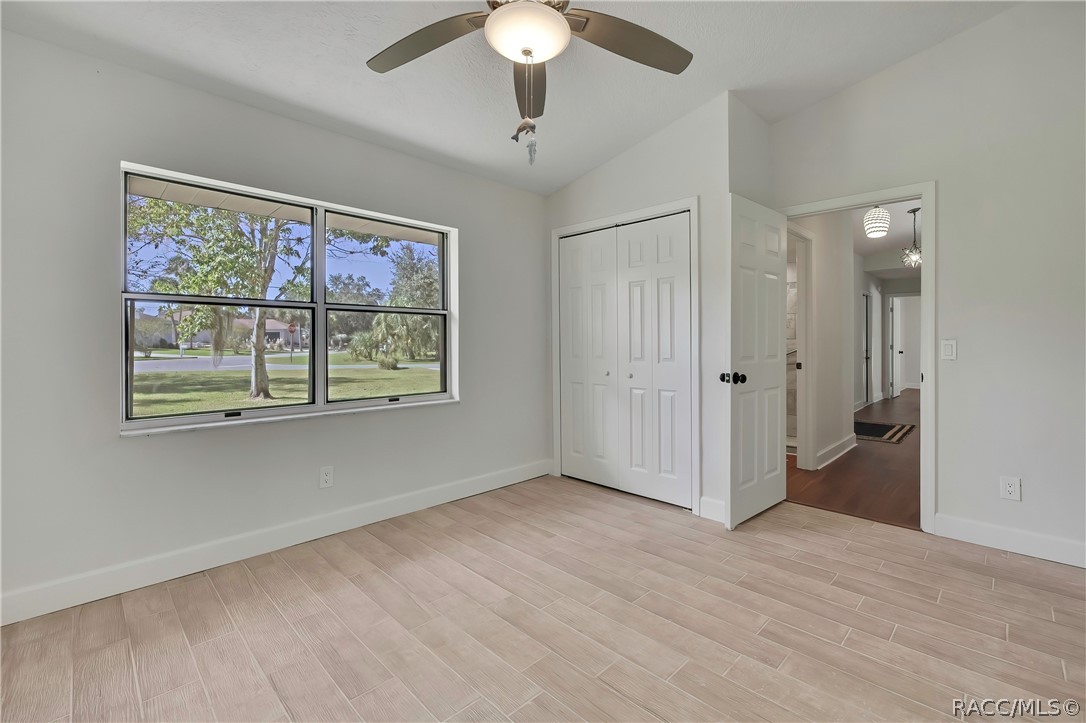 Empty bedroom: light plank tile floor, a large double window looking onto the street, front lawn, trees and neighbouring houses, white double closet doors in the centre, an open doorway at the right to a hallway with a dark wood floor and pendant lights, a ceiling fan with a dome light, and white walls. No furniture.