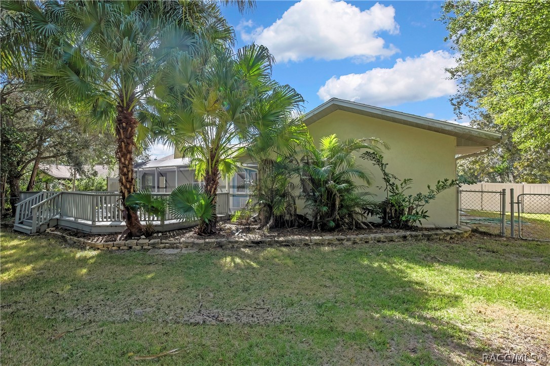 Rear of the house from the lawn: light stucco walls, a shingle roof, a stucco chimney with a metal cap, a screen enclosure with blue doors, a grey wood deck with steps and railing, palm trees, a fenced lawn in the foreground, and neighbouring houses and chain-link and vinyl fences at the sides.