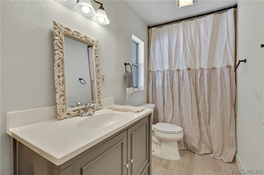 Primary bathroom, other angle: grey vanity with a white counter and an ornate white-framed mirror, a chrome faucet, a folded towel on the counter, a white toilet, a tub/shower with a white-and-striped curtain on a black rod, a narrow window, light plank tile floor, a 3-light fixture at the top of the frame, and a ceiling vent.