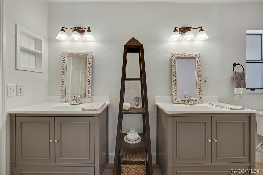 Primary bathroom with dual vanities: two grey vanity cabinets with white counters and chrome faucets, two ornate white-framed mirrors, a 3-light fixture above each mirror, a dark ladder shelf between the sinks with a bust, a white flower, a vase and a basket below, folded towels on each counter, a recessed wall niche at the left, a towel ring with a grey-and-white towel and a window at the right, and the edge of a toilet at the far right.