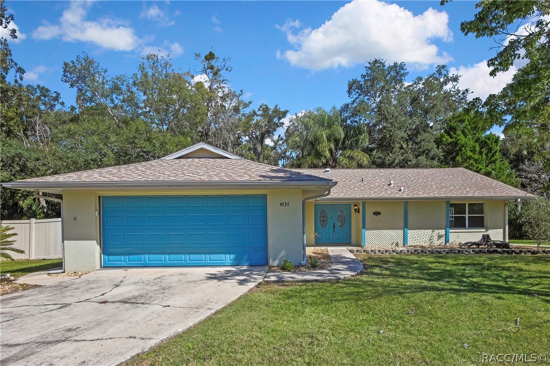 Straight-on front of the single-storey house: light cream stucco walls, grey shingle roof with a gable over the garage, wide blue panelled garage door, house number 4737 on the wall beside the garage, blue double entry doors with oval decorative glass, a wall light beside the doors, a lattice wall section and a large front window, concrete driveway and a curved concrete walkway to the entry, a stone-edged planting bed, green lawn, trees and palms, white vinyl fence at the left edge, blue sky
