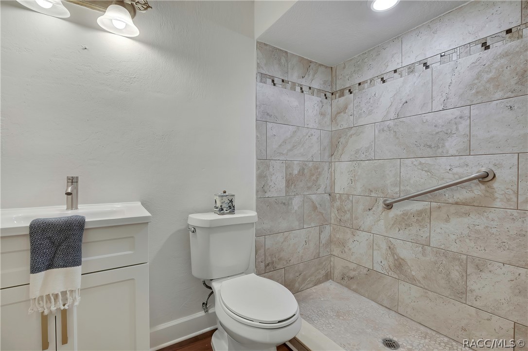 Bathroom: white vanity with a modern chrome faucet, a navy-and-white towel hanging over the vanity, a white toilet with a patterned container on the tank, a walk-in shower with stone-look tile walls, a mosaic accent strip, a grab bar and a pebble floor with a drain, recessed ceiling lights, and a 2-light fixture at the top left. This is the single-sink hall bath, not the primary bath.