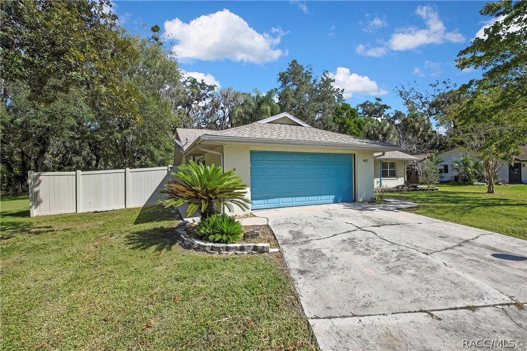 Front-garage angle: blue panelled garage door, house number 4737 on the wall beside it, wide concrete driveway in the foreground, white vinyl fence at the left, a palm plant in a stone-edged bed in front of the garage, green lawn, a neighbouring house at the right, trees, blue sky with white clouds.