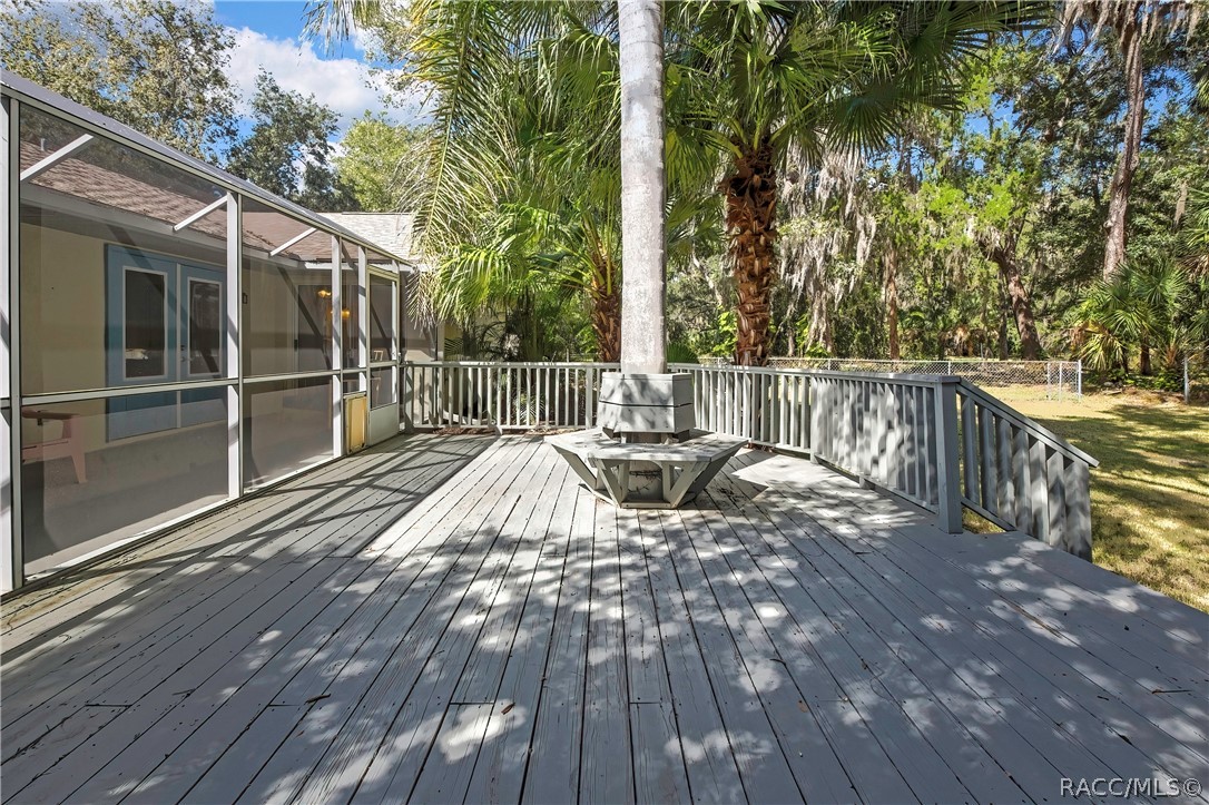 Wood deck: grey wood deck planks, railing, a built-in bench around a palm tree trunk, steps down to a fenced lawn, palm trees, oaks with Spanish moss, and a chain-link fence in the background.