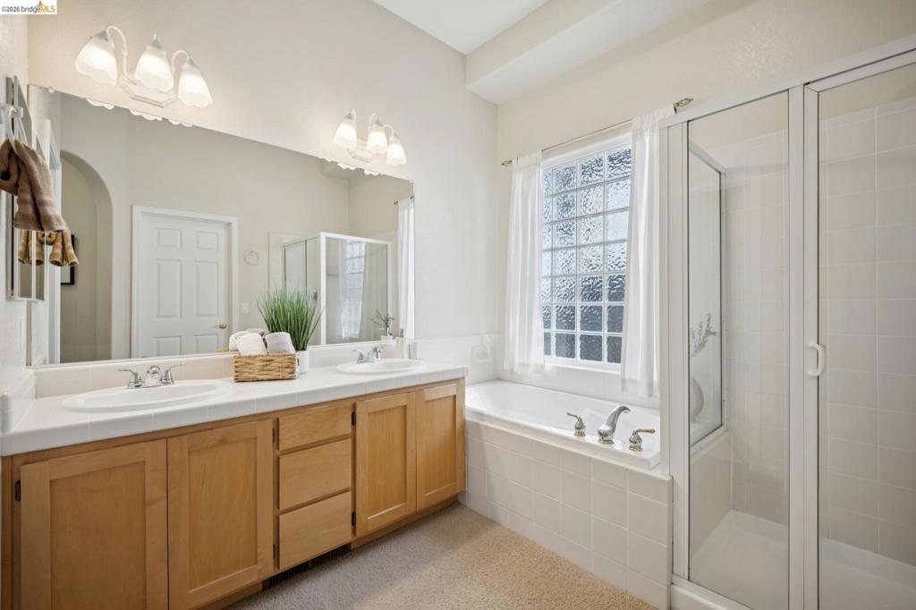 Dual-sink vanity with light-tone wood cabinetry, porcelain-tiled tub deck, glass-enclosed shower, and textured glass window