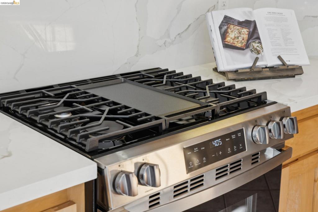 Stainless steel gas range with a central griddle, heavy-duty iron burner grates, digital touch controls, and light-toned cabinetry complemented by white stone-finish slab countertops with veining