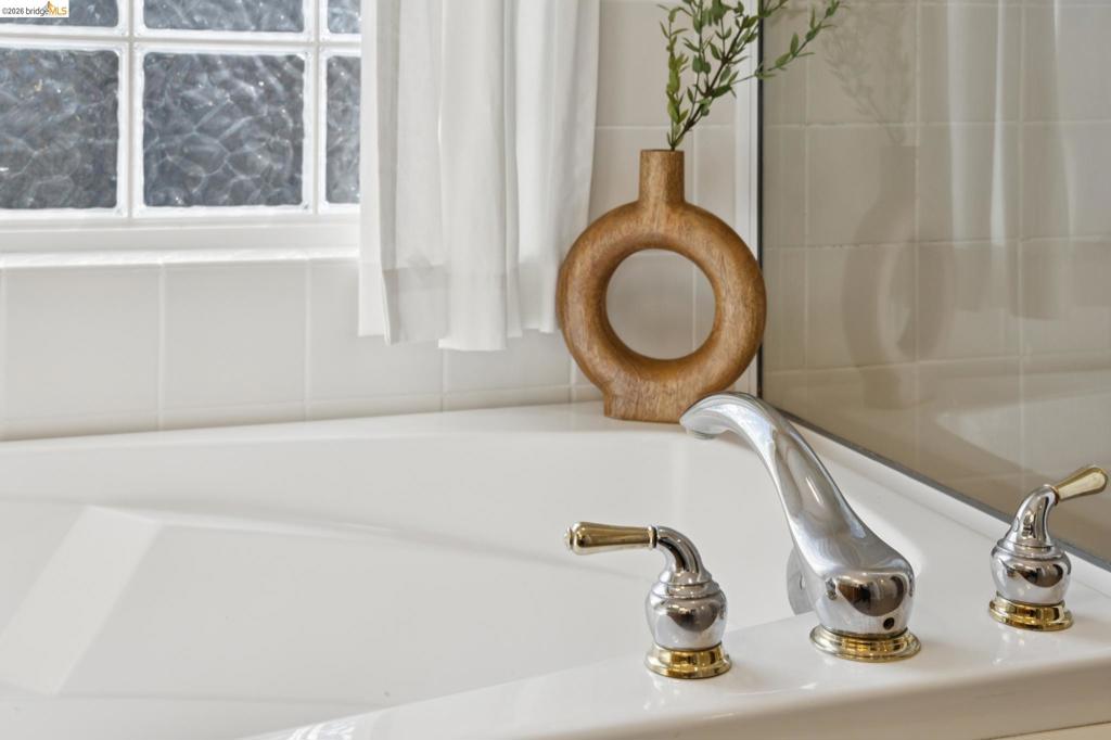 Soaking tub featuring a white acrylic finish, deck-mounted chrome faucet with gold-tone accents, square tile surround, and an integrated glass block window