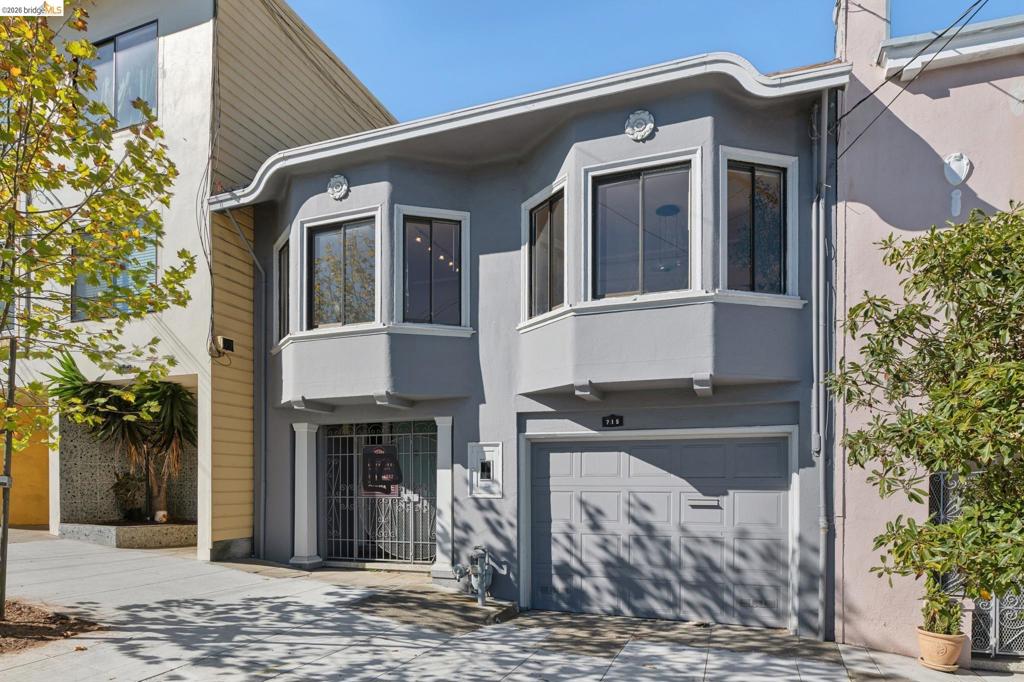 Exterior facade featuring a smooth stucco finish, dual upper-level bay windows, decorative crown medallions, a metal security gate, and an integrated single-car garage