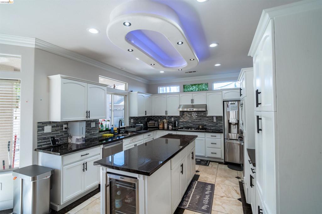 Kitchen featuring custom white cabinetry, a center island with built-in wine refrigeration, dark stone countertops, a tiled backsplash, and an elliptical recessed ceiling feature with integrated lighting
