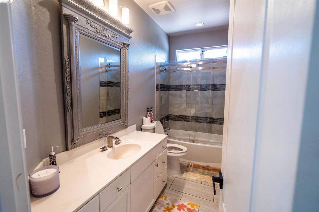 Bathroom featuring a vanity with a white countertop and oval sink, a wall-mounted decorative frame mirror, recessed lighting, and a bathtub with a glass shower enclosure and tiled surround