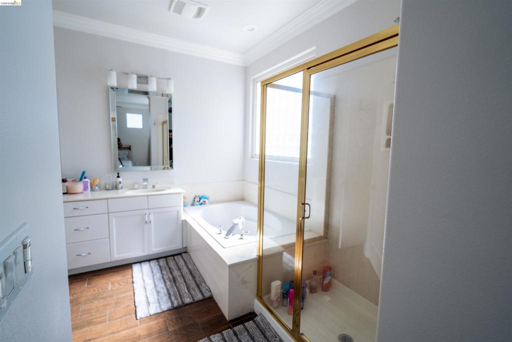Bathroom featuring white cabinetry, built-in soaking tub with a stone-finish surround, glass-enclosed shower with gold-tone frame, and wood-finish flooring