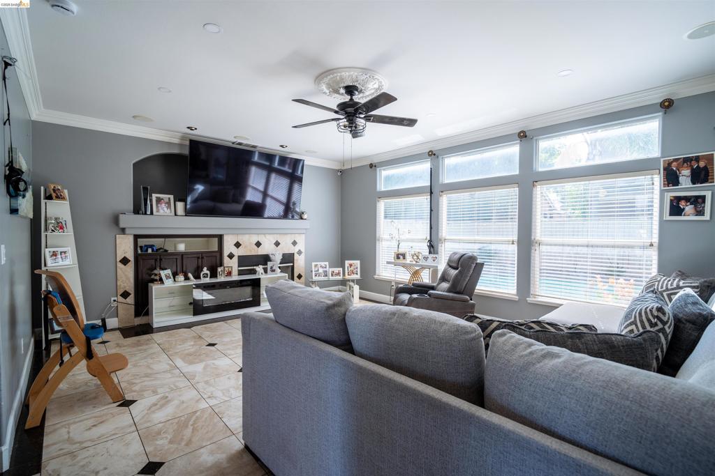 Living area featuring oversized floor tiles with decorative inlay, a fireplace with integrated cabinetry, crown molding, a central ceiling fan with medallion detail, and a wall of multi-pane windows
