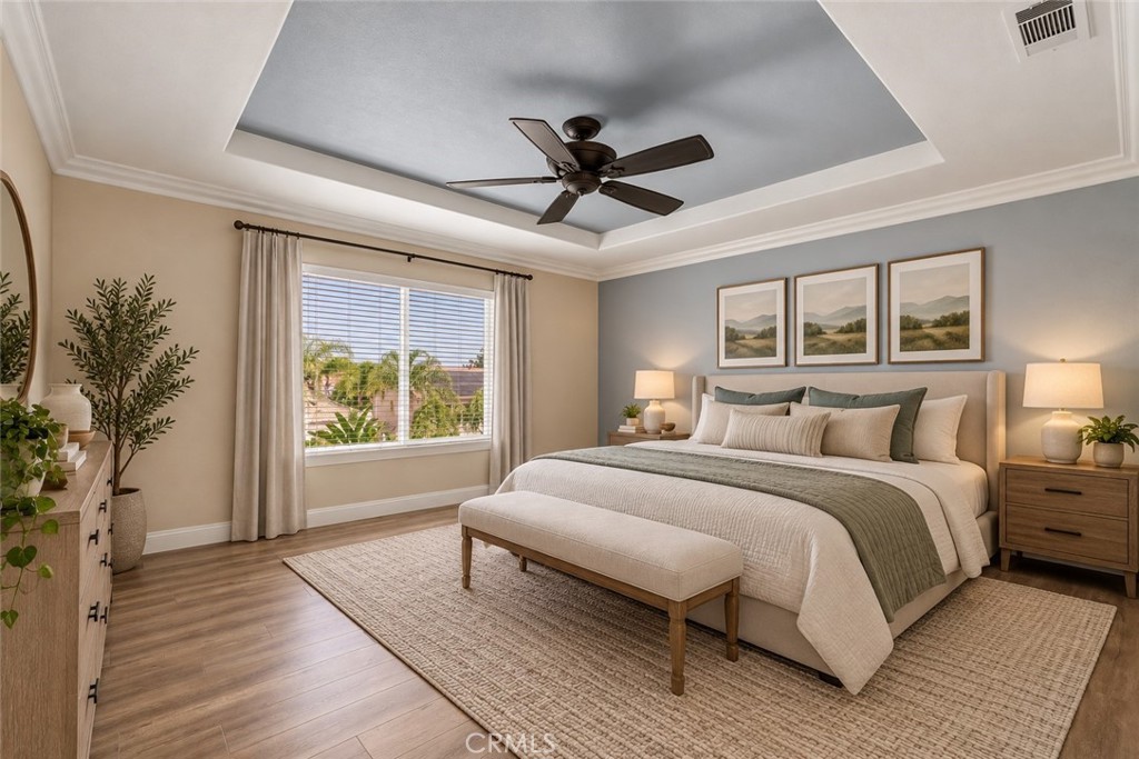 Beautiful Primary Suite Featuring A Decorative Tray Ceiling With Crown Molding, Ceiling Fan, Attractive Wood-Look Flooring, And A Large Window Providing Abundant Natural Light. Digitally Staged