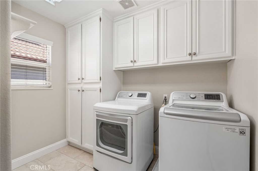 Convenient Indoor Laundry Room Featuring A Side-By-Side Washer And Dryer, Abundant Built-In Cabinetry For Storage, Tile Flooring, And A Window Providing Natural Light.