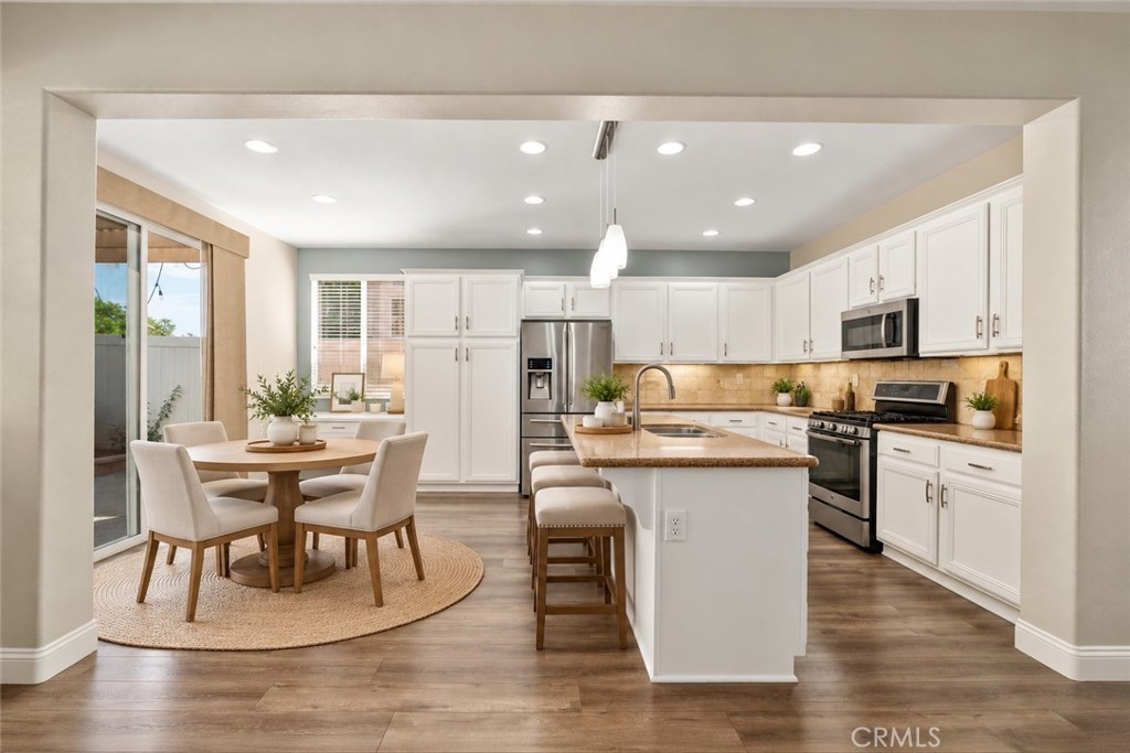 Spacious Kitchen And Adjacent Dining Area Featuring White Cabinetry, Stainless Steel Appliances, A Large Center Island, Recessed Lighting, And Sliding Glass Door Access To The Backyard. Digitally Staged