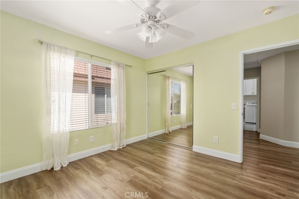 Another View Of The Bedroom Highlighting The Mirrored Closet Doors, Ceiling Fan, Wood-Look Flooring, And Large Window With Soft Window Treatments.