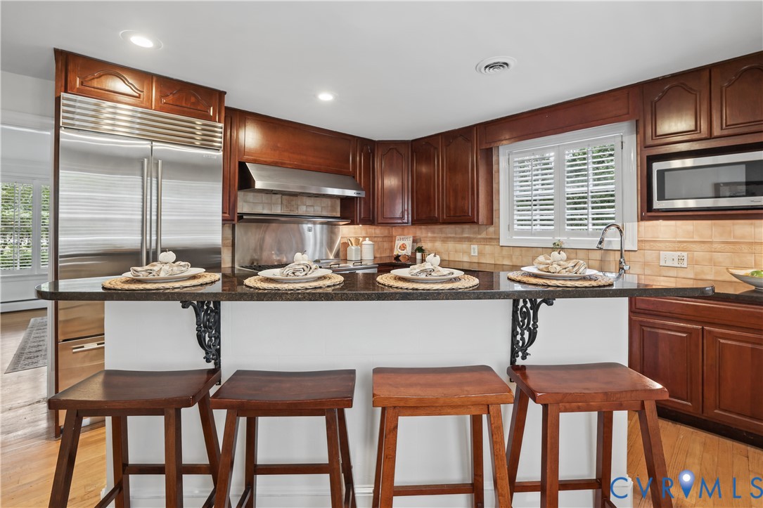 Kitchen featuring stained wood cabinetry, stone countertops with an integrated breakfast bar, stainless steel refrigerator, built-in microwave, and subway tile backsplash