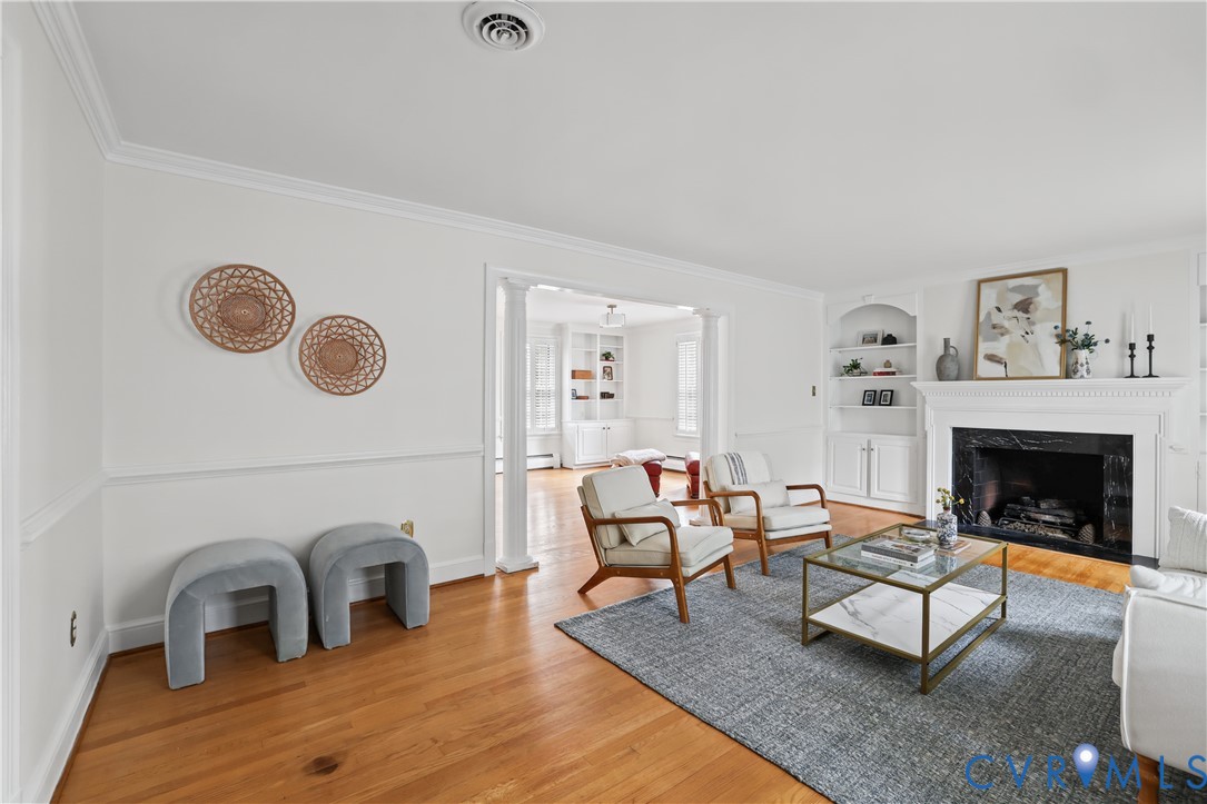 Living area featuring wood-finish flooring, a fireplace with a stone-surround and mantle, built-in shelving, decorative crown molding, and architectural columns