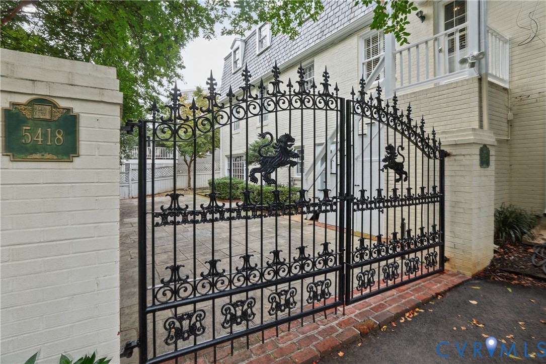 Ornate black wrought iron entry gates featuring lion crest motifs, set between brick pillars, leading to a stone-paved courtyard and a brick exterior with a mansard roof