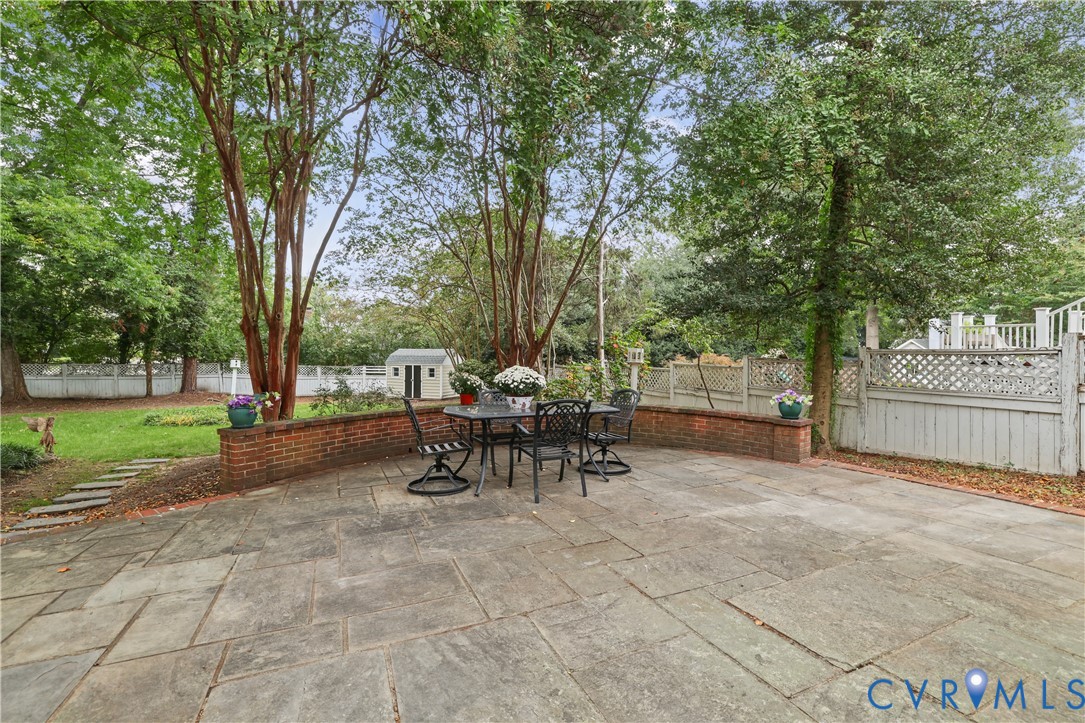 Flagstone patio featuring a low-profile brick retaining wall, mature trees, lattice-style fencing, and a detached storage structure