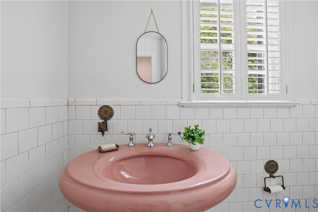 Wall-mounted ceramic pedestal sink in a curved silhouette, white square tile wainscoting, and window with white plantation shutters